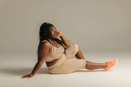 Téléchargez les photos : Belle femme africaine taille plus en vêtements de sport regardant confiant tout en étant assis sur fond de studio - en image libre de droit