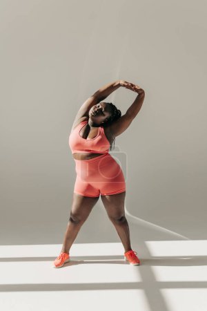 Téléchargez les photos : Pleine longueur de confiance africaine plus la femme de taille dans les vêtements de sport faire des exercices d'étirement dans le studio de fitness - en image libre de droit