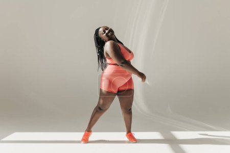 Foto de Longitud completa de la mujer africana feliz más tamaño en ropa deportiva que irradia amor propio mientras está de pie en el estudio de fitness - Imagen libre de derechos