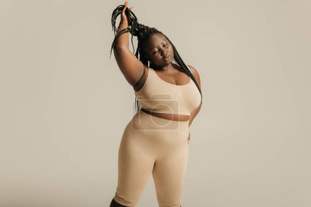 Téléchargez les photos : Belle africaine taille plus femme en sous-vêtements rayonnant d'amour-propre tout en ajustant ses dreadlocs sur fond de studio - en image libre de droit