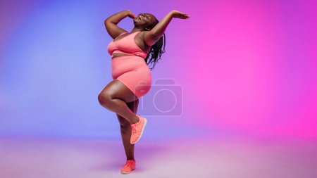 Téléchargez les photos : Pleine longueur de femme africaine voluptueuse joyeuse en vêtements de sport dansant et souriant sur fond coloré - en image libre de droit