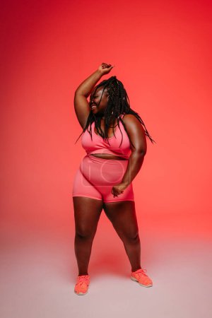 Foto de Mujer Africana feliz de talla grande en ropa deportiva bailando e irradiando amor propio contra un fondo vibrante - Imagen libre de derechos