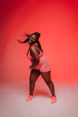 Photo for Curvy young African woman in sportswear dancing and radiating self-love against vibrant background - Royalty Free Image