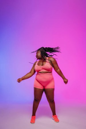 Foto de Hermosa mujer africana de talla grande en ropa deportiva bailando y temblando dreadlocs contra fondo colorido - Imagen libre de derechos