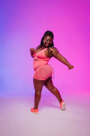 Foto de Alegre mujer africana de talla grande en ropa deportiva bailando e irradiando amor propio contra un fondo colorido - Imagen libre de derechos