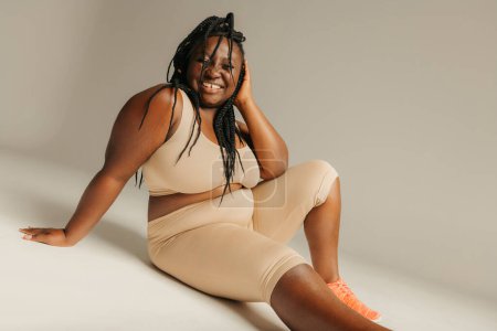 Téléchargez les photos : Happy plus taille femme africaine en vêtements de sport rayonnant d'amour-propre tout en étant assis sur fond de studio - en image libre de droit