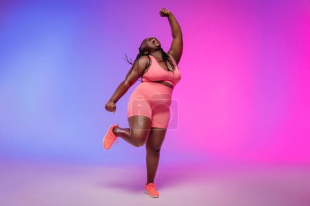 Foto de Longitud completa de la alegre mujer con curvas en ropa deportiva bailando y sonriendo sobre un fondo colorido - Imagen libre de derechos