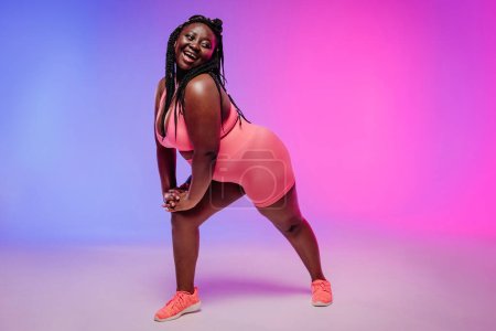 Téléchargez les photos : Jeune femme africaine voluptueuse dans l'exercice de vêtements de sport et regardant heureux sur fond coloré - en image libre de droit