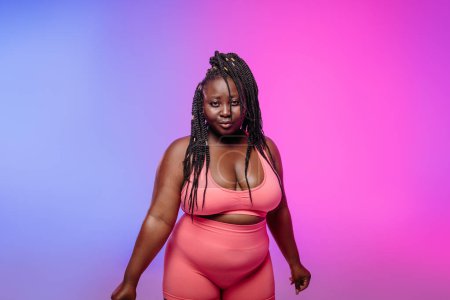 Foto de Confiado africano más mujer de tamaño en ropa deportiva mirando a la cámara contra el fondo colorido - Imagen libre de derechos