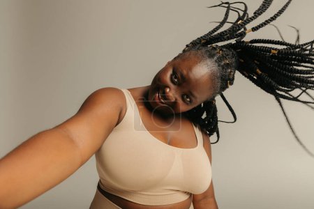 Téléchargez les photos : Joyeuse femme africaine taille plus avec dreadlocks faire selfie tout en se tenant debout sur fond de studio - en image libre de droit