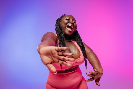 Foto de Hermosa mujer africana de talla grande en ropa deportiva haciendo gestos y sonriendo sobre un fondo vibrante - Imagen libre de derechos