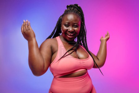 Foto de Hermosa mujer africana con curvas en ropa deportiva haciendo gestos y sonriendo sobre un fondo vibrante - Imagen libre de derechos