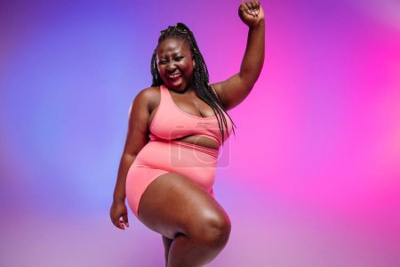 Foto de Hermosa mujer de talla grande en ropa deportiva irradiando amor a su cuerpo mientras baila sobre un fondo vibrante - Imagen libre de derechos