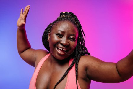 Téléchargez les photos : Belle femme africaine voluptueuse en vêtements de sport gestuelle et souriante sur fond vibrant - en image libre de droit