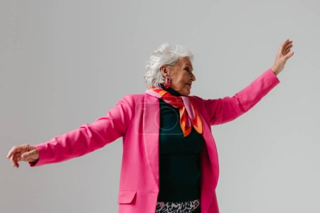 Téléchargez les photos : Femme âgée élégante en vêtements à la mode rayonnant d'amour-propre tout en dansant sur fond gris - en image libre de droit