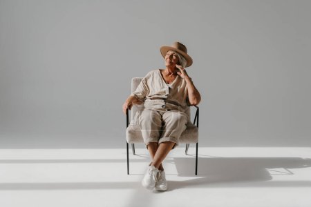 Téléchargez les photos : Femme âgée cheveux gris détendue dans un chapeau élégant assis dans une chaise confortable au studio ensoleillé - en image libre de droit