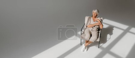 Téléchargez les photos : Vue du dessus des cheveux gris calme femme âgée relaxant dans une chaise confortable dans un studio ensoleillé - en image libre de droit