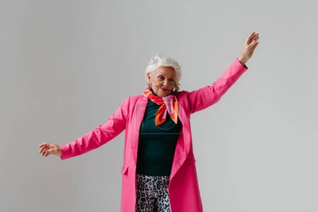 Téléchargez les photos : Femme âgée élégante en vêtements à la mode rayonnant d'amour-propre tout en dansant sur fond gris - en image libre de droit