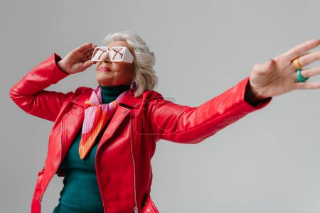 Téléchargez les photos : Cool femme âgée en veste en cuir rouge et lunettes de vue tendance gestuelle tout en dansant sur fond gris - en image libre de droit