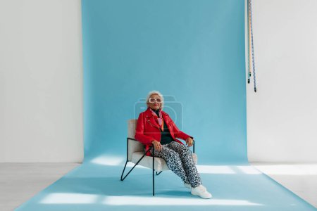 Téléchargez les photos : Cool femme âgée en tenue à la mode assis dans une chaise confortable sur fond bleu dans le studio photo - en image libre de droit