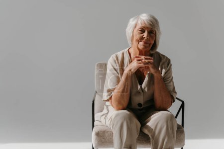 Téléchargez les photos : Cheveux gris détendu femme âgée assise dans une chaise confortable et regardant la caméra dans un studio ensoleillé - en image libre de droit