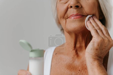 Téléchargez les photos : Gros plan de la femme âgée nettoyant le visage avec une éponge de coton sur fond gris - en image libre de droit