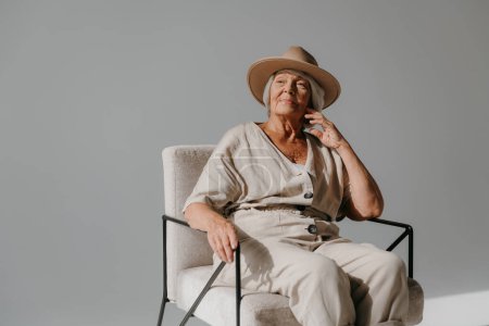 Téléchargez les photos : Elégante femme âgée aux cheveux gris en chapeau relaxant dans une chaise confortable au studio ensoleillé - en image libre de droit