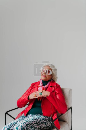 Téléchargez les photos : Cool femme âgée en tenue à la mode assis dans une chaise confortable sur fond gris - en image libre de droit