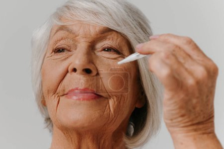 Foto de Mujer mayor segura usando suero cosmético mientras disfruta del tratamiento de belleza contra el fondo gris - Imagen libre de derechos