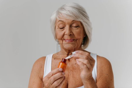 Téléchargez les photos : Femme âgée confiante utilisant un sérum cosmétique tout en profitant d'un traitement de beauté sur fond gris - en image libre de droit