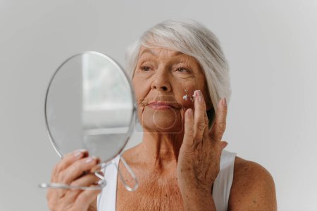 Foto de Mujer mayor de confianza usando espejo mientras se aplica crema anti-edad en la cara contra el fondo gris - Imagen libre de derechos