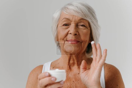 Téléchargez les photos : Femme âgée confiante utilisant la crème cosmétique tout en se tenant debout sur fond gris - en image libre de droit