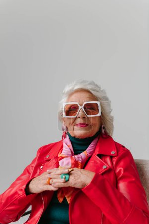 Téléchargez les photos : Cool femme âgée en tenue à la mode assis dans une chaise confortable sur fond gris - en image libre de droit