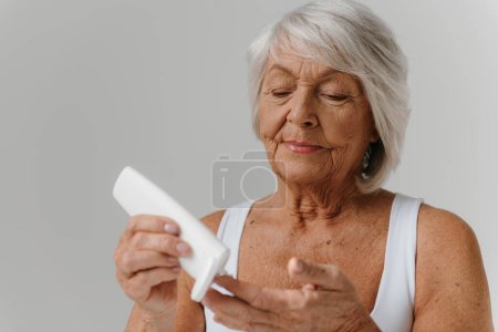 Téléchargez les photos : Femme âgée confiante versant de la crème cosmétique sur les doigts sur fond gris - en image libre de droit
