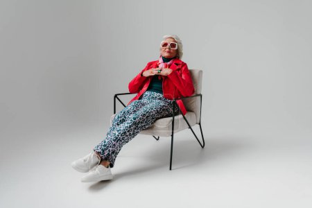 Téléchargez les photos : Cool femme âgée en vêtements à la mode assis dans une chaise confortable sur fond gris - en image libre de droit