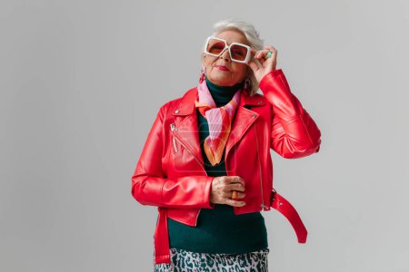 Téléchargez les photos : Femme senior à la mode en veste en cuir rouge ajustant les lunettes à la mode sur fond gris - en image libre de droit