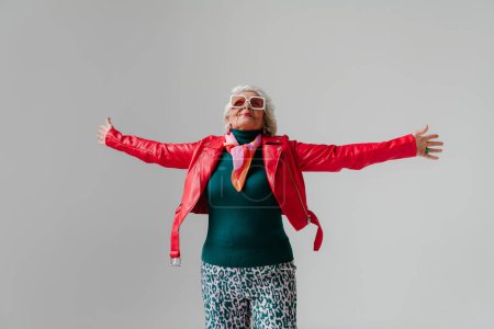 Photo for Fashionable senior woman in red leather jacket and trendy eyeglasses stretching out hands on grey background - Royalty Free Image