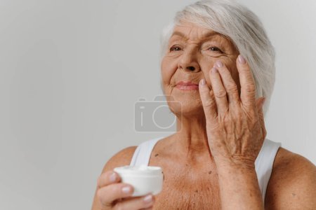 Téléchargez les photos : Femme âgée confiante appliquant une crème anti-âge sur le visage sur fond gris - en image libre de droit