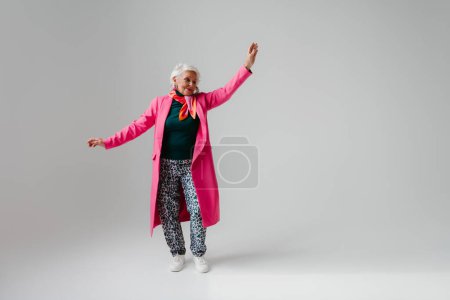 Téléchargez les photos : Pleine longueur de femme âgée élégante en vêtements à la mode rayonnant d'amour-propre tout en dansant sur fond gris - en image libre de droit