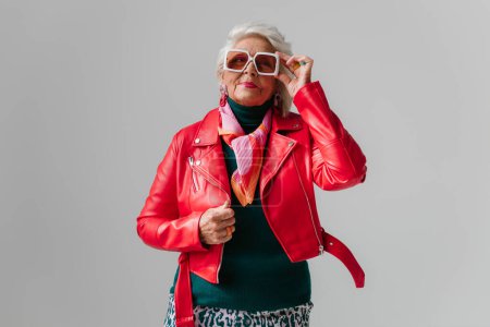Téléchargez les photos : Femme senior à la mode en veste en cuir rouge et lunettes tendance à la recherche cool sur fond gris - en image libre de droit