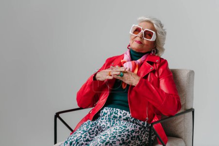 Téléchargez les photos : Femme senior à la mode en veste en cuir rouge assise dans une chaise confortable sur fond gris - en image libre de droit