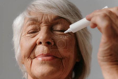 Téléchargez les photos : Gros plan d'une femme chevelue grise qui applique de la crème anti-âge sous les yeux sur fond gris - en image libre de droit
