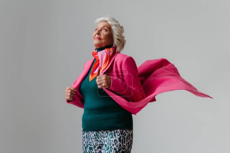 Foto de Elegante anciana con aspecto de súper héroe mientras usa abrigo rosa sobre fondo gris - Imagen libre de derechos