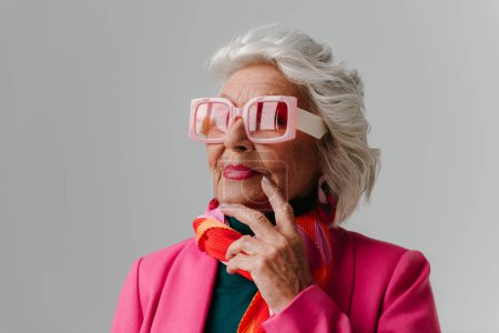Téléchargez les photos : Élégante femme âgée portant des lunettes de vue de style rétro à la mode et rayonnant d'amour-propre sur fond gris - en image libre de droit