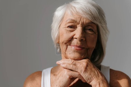 Téléchargez les photos : Portrait de femme âgée souriante penchée la tête sur les mains et souriant sur fond gris - en image libre de droit
