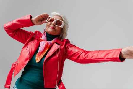 Photo for Fashionable elderly woman in leather jacket and trendy eyeglasses gesturing while dancing on grey background - Royalty Free Image