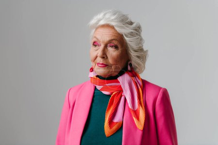Foto de Elegante anciana con maquillaje vestida con ropa de moda e irradiando amor propio sobre fondo gris - Imagen libre de derechos