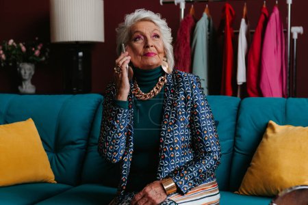 Photo for Elegant elderly gray hair woman in fashionable wear talking on phone while sitting on the couch at home - Royalty Free Image