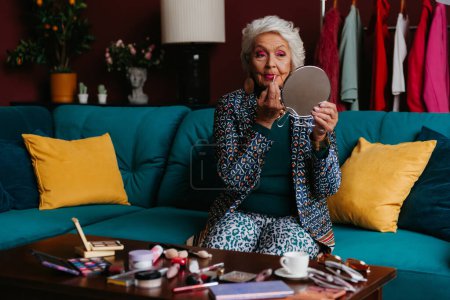 Téléchargez les photos : Femme âgée confiante dans le port à la mode faisant maquillage tout en étant assis sur le canapé à la maison - en image libre de droit