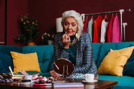 Foto de Mujer de edad avanzada con estilo en ropa de moda aplicando lápiz labial mientras está sentado en el sofá en casa - Imagen libre de derechos